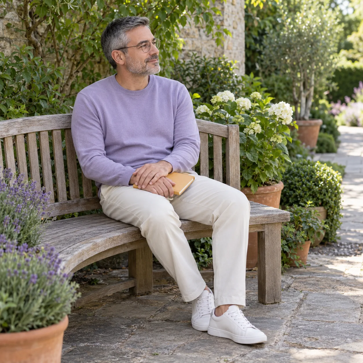 A fictional adult man in a lavender top sits on a wooden garden bench with both hands over a closed ochre notebook in his lap.