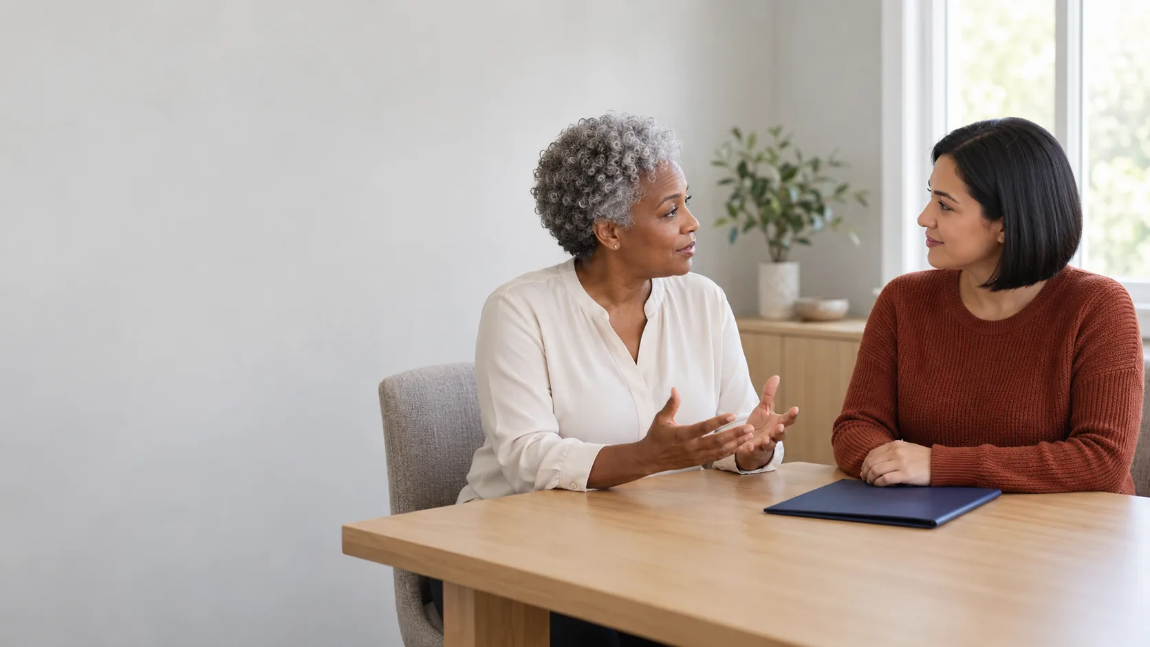 Desktop illustration: Two women talking across a pale wooden table beside a closed navy folder, with pale wall space on the left. Mobile illustration: A woman in a teal cardigan looking at an open notebook and holding a pen at a wooden home table beneath a pale upper wall.