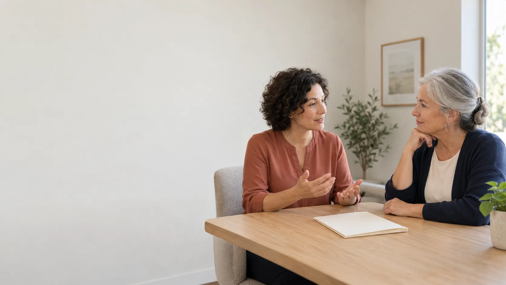 Desktop illustration: Two adult women talking across a wooden table beside a closed cream folder, with pale wall space on the left. Mobile illustration: Adult woman holding a pen over an open blank notebook at a wooden table, with pale wall space above.