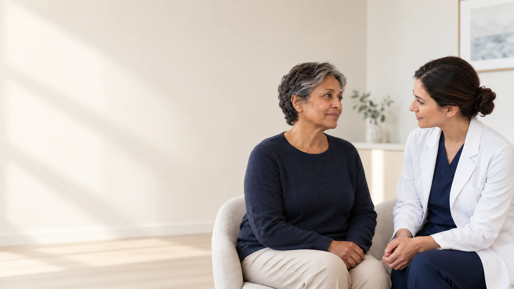 A fictional mature adult visitor speaks with a fictional woman clinician in a calm consultation room, with the visitor's natural cheek, jawline and upper neck visible.