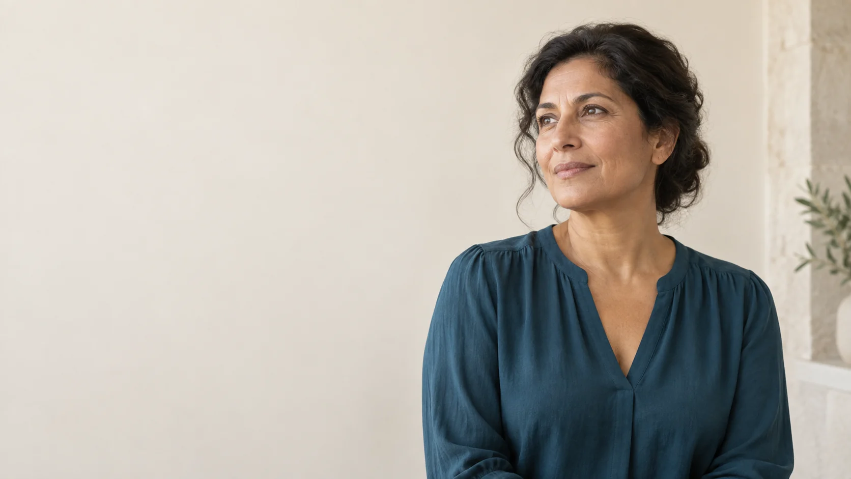 A fictional adult woman with dark wavy hair and a deep-teal blouse in a softly lit ivory room.