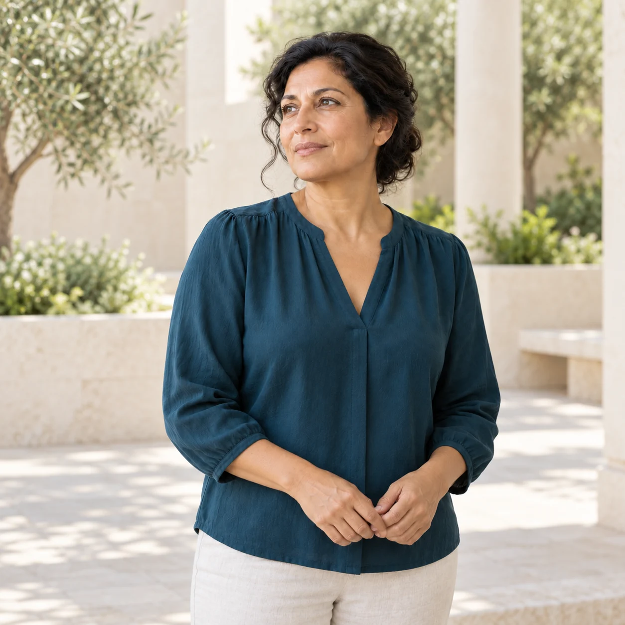The same fictional woman in a deep-teal blouse stands in a softly lit pale-stone courtyard.