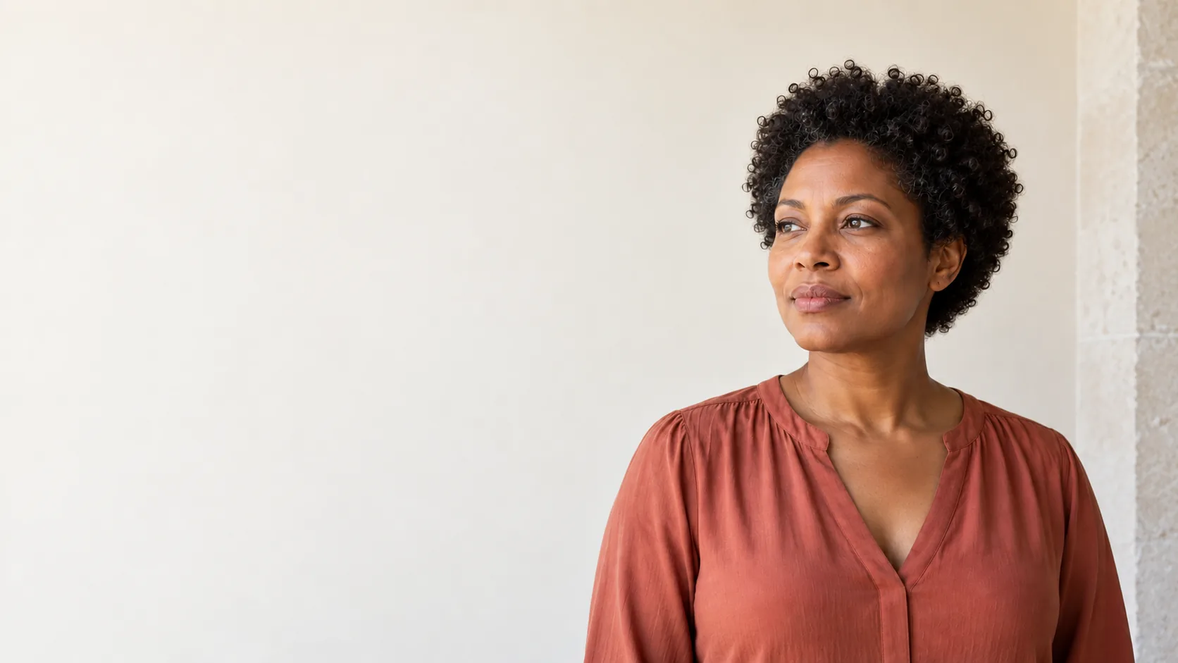 A fictional adult woman with short natural curls and a muted terracotta blouse stands beside an open ivory wall.