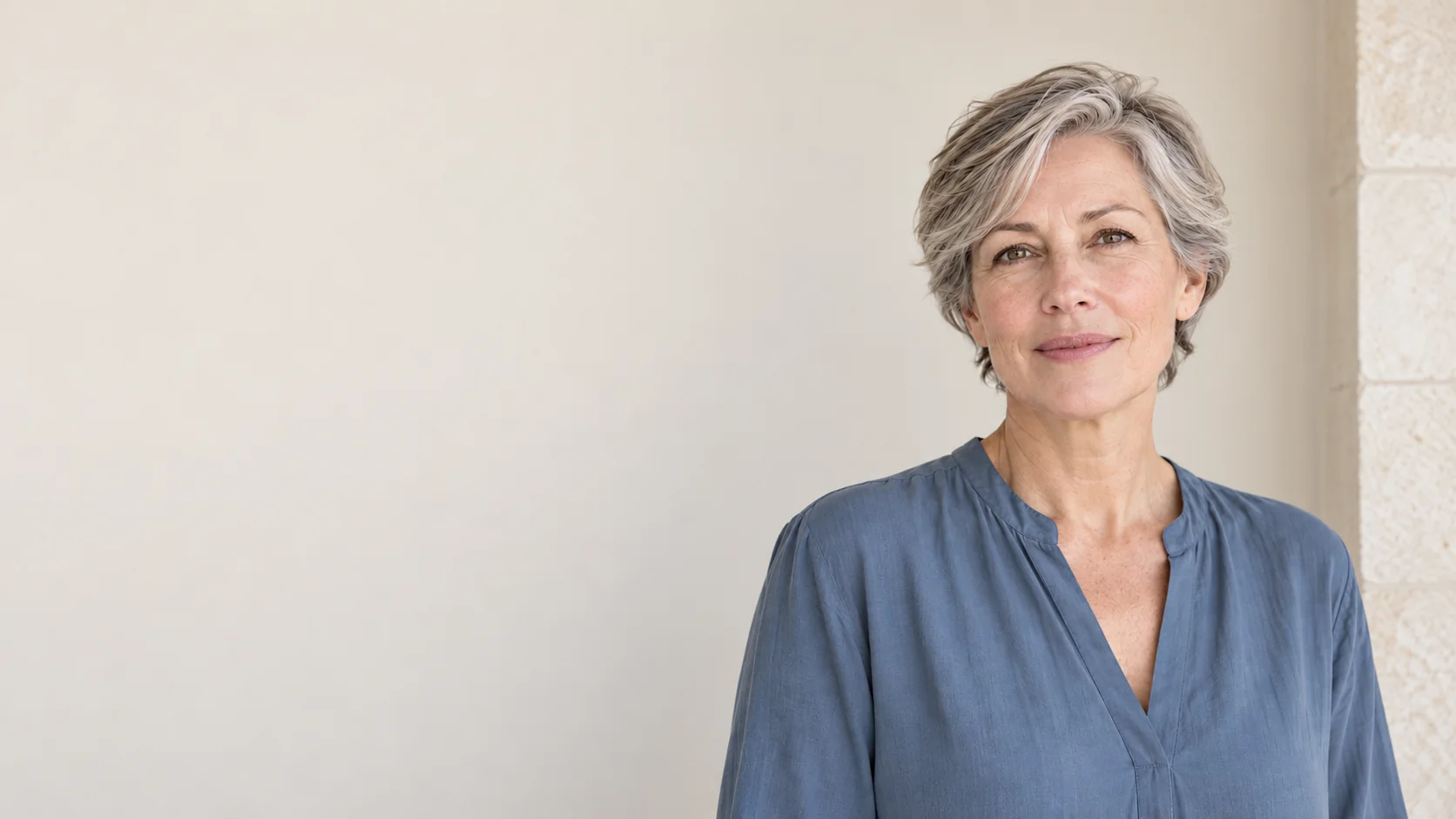 A fictional silver-haired adult woman in a slate-blue blouse stands against a softly lit ivory wall.