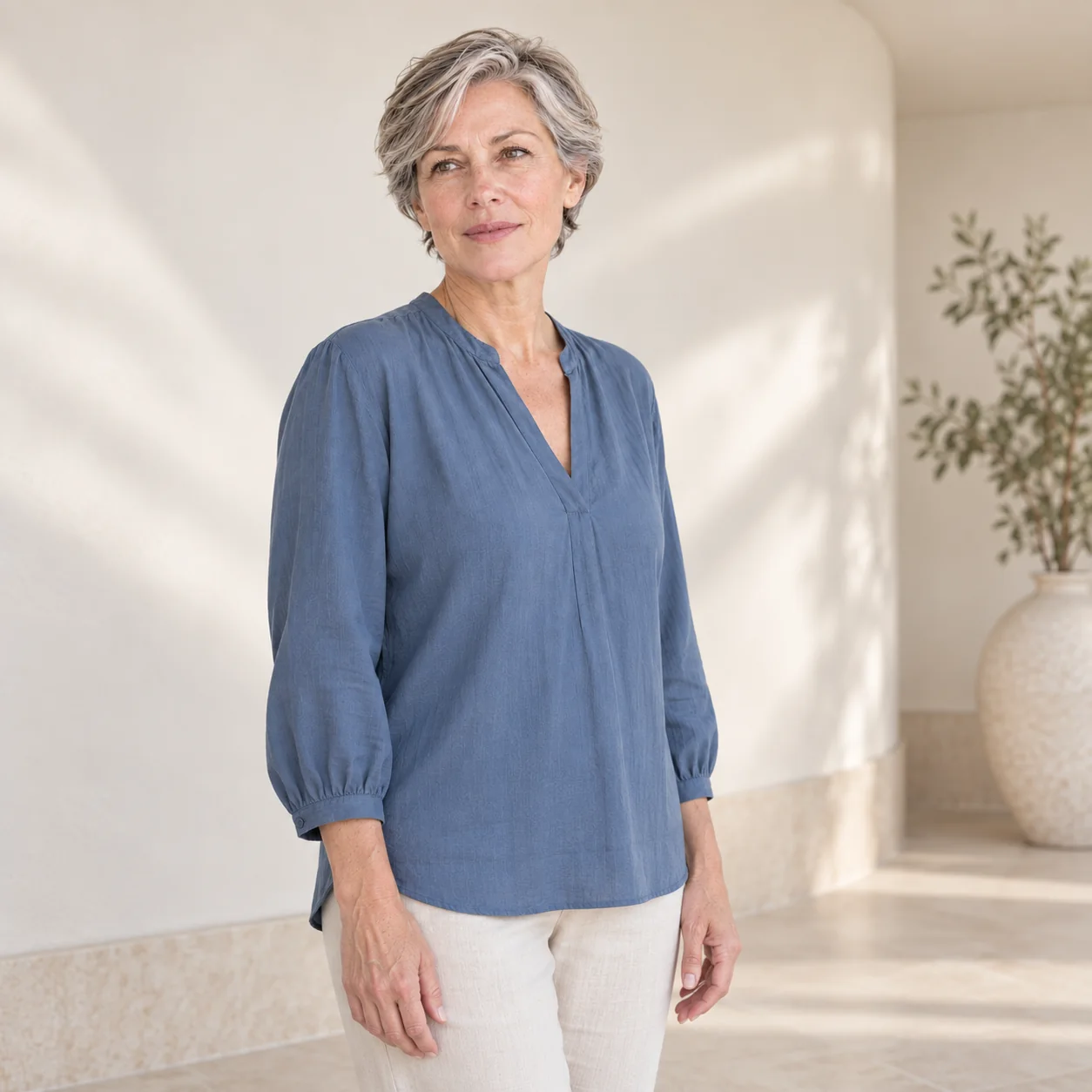 The same fictional silver-haired woman in a slate-blue blouse stands in a softly lit ivory room with a curved plaster wall.