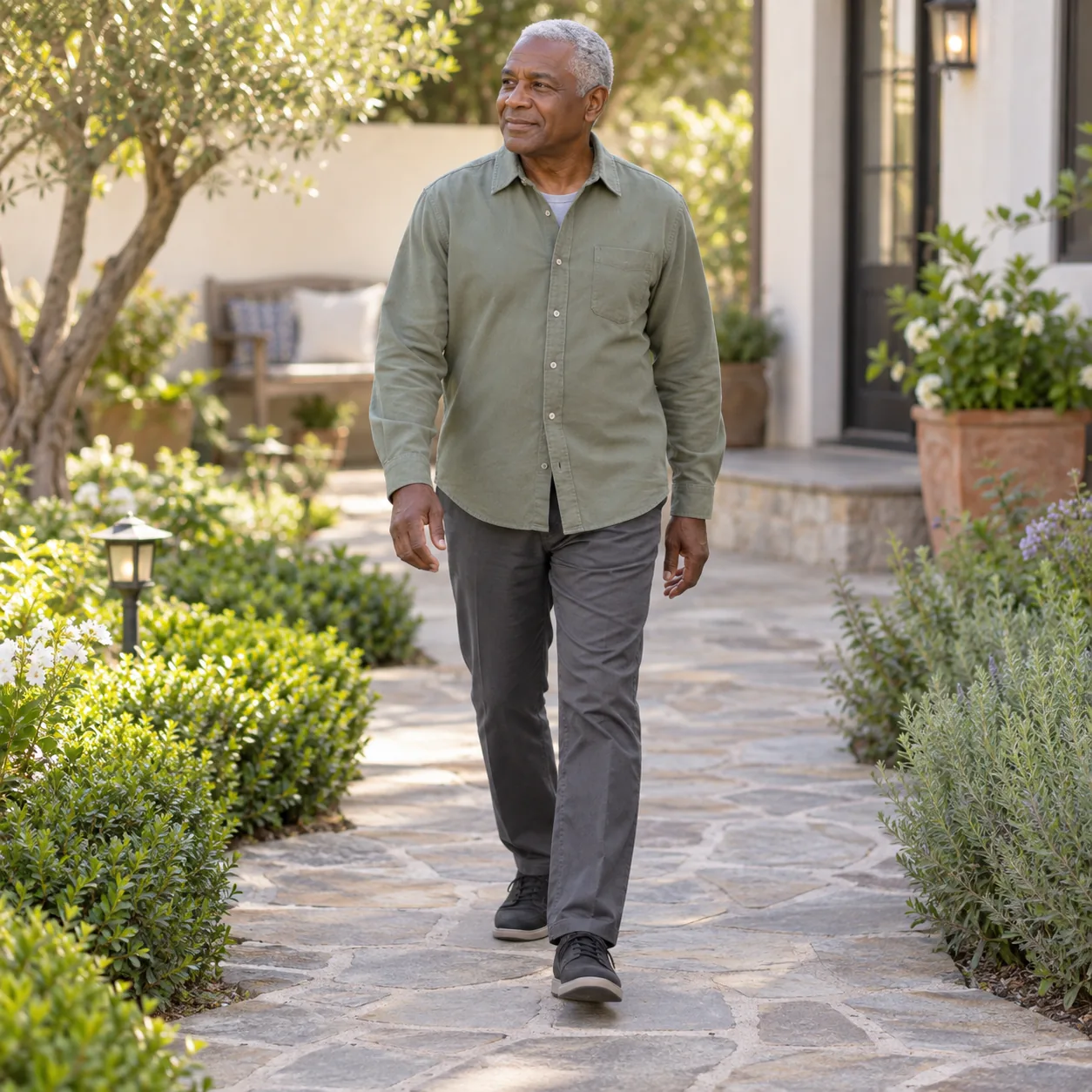 An older man in a sage-green shirt walks along a stone garden path.