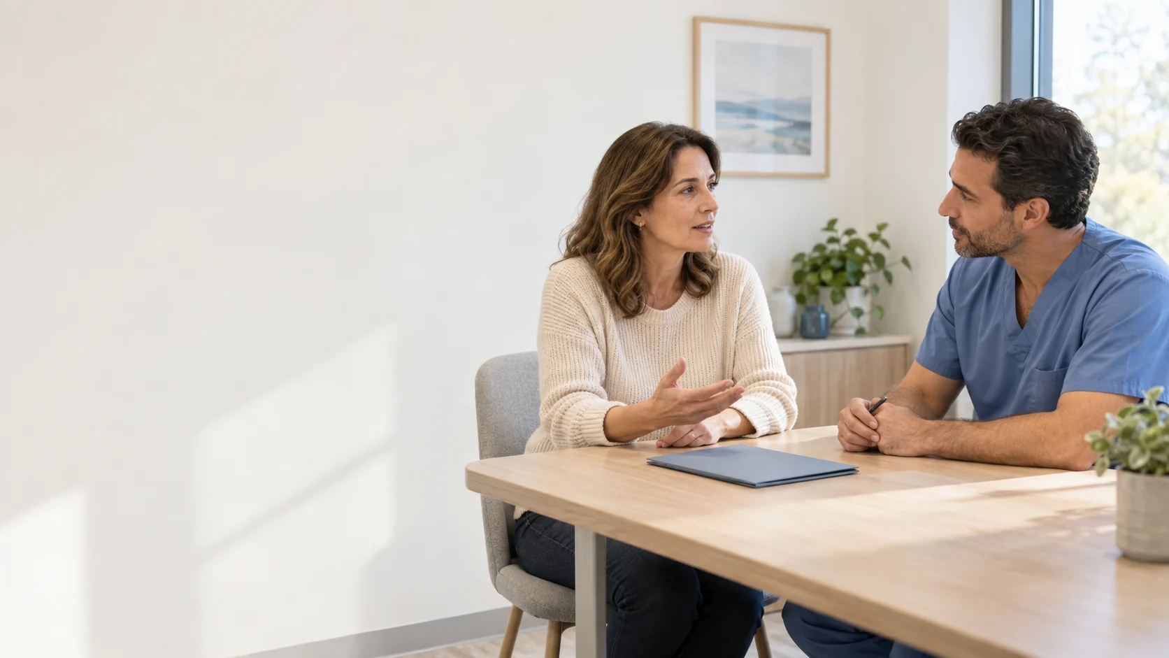 Two adults talking across a table in a consultation room; one wears blue clinical attire.